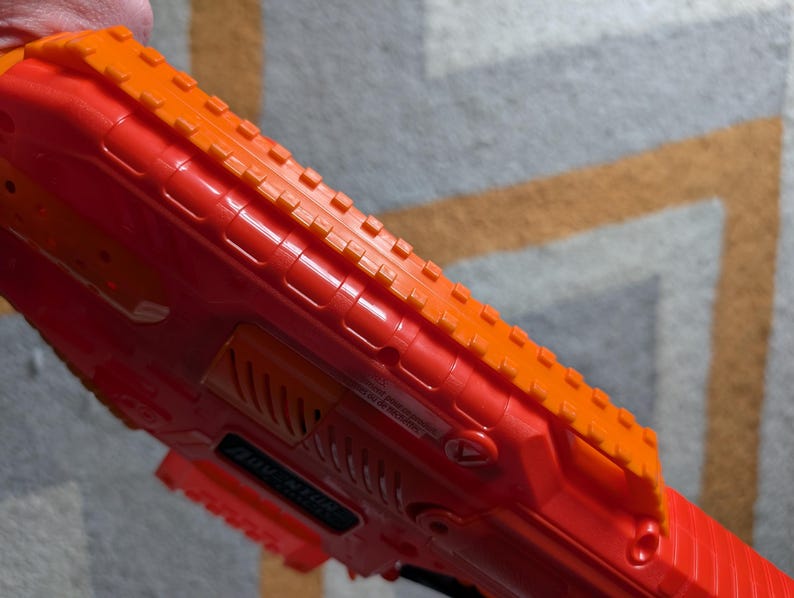 May include: Close-up of a red and orange toy gun. The toy gun has a textured orange top rail and a red body. The gun has a series of ridges and grooves along its surface. The toy gun is held by a person.