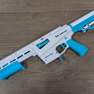 May include: A white and bright blue toy blaster with an orange tip. The side of the blaster features the text GAME FACE and TRION. It includes a long barrel with a rail system, a blue grip, a blue trigger, and an adjustable white and blue stock.