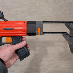 May include: An orange and gray toy gun with the text "SPECTRUM" on the side. The gun has a black grip, a gray stock, and an orange trigger. The gun is held by a person.