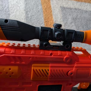 May include: A red and orange toy gun with a black and orange scope attached. The scope has a cylindrical orange end and a black body. The gun has orange accents and a textured surface.