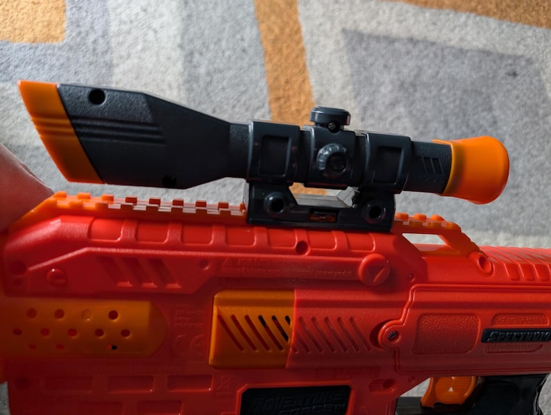 May include: A red and orange toy gun with a black and orange scope attached. The scope has a cylindrical orange end and a black body. The gun has orange accents and a textured surface.