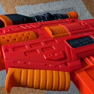 May include: A red and orange Adventure Force toy gun with a black scope. The toy gun has an orange handle and accents. The text "Adventure Force" is visible on the side of the gun. The toy gun is lying on a patterned surface.
