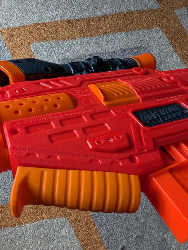 May include: A red and orange Adventure Force toy gun with a black scope. The toy gun has an orange handle and accents. The text "Adventure Force" is visible on the side of the gun. The toy gun is lying on a patterned surface.