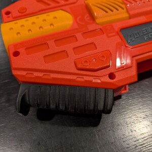 May include: Close-up of a red and orange Adventure Force toy gun. The toy gun has a black handle and the words "Adventure Force" are visible on the side. The gun is made of plastic.