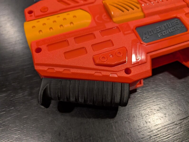 May include: Close-up of a red and orange Adventure Force toy gun. The toy gun has a black handle and the words "Adventure Force" are visible on the side. The gun is made of plastic.