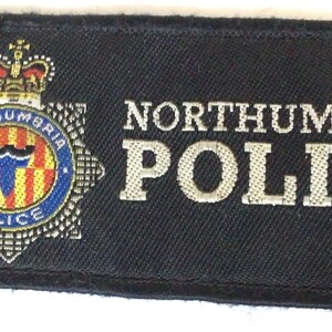 May include: A rectangular black patch with the words "NORTHUMBRIA POLICE" in white lettering. The patch features a detailed emblem with a crown and the words "NORTHUMBRIA POLICE" in a circular design.