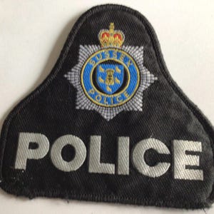 May include: A black police patch with the word "POLICE" in silver lettering. The patch features a blue and silver emblem with a crown and the words "SUSSEX POLICE". The emblem is at the top of the patch.