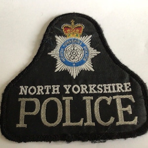 Könnte beinhalten: Ein schwarzer, gestickter Aufnäher in Glockenform. Der Aufnäher zeigt oben das Emblem der North Yorkshire Police mit einer Krone und einer weißen Rose, darunter der in weißen Buchstaben gestickte Text NORTH YORKSHIRE POLICE.