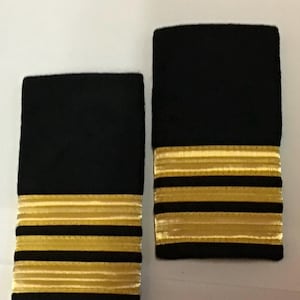 Könnte beinhalten: Zwei schwarze Schulterstücke mit goldenen Streifen. Die Schulterstücke sind rechteckig, mit einem einfarbigen schwarzen oberen Teil und mehreren goldenen Streifen am unteren Rand. Die Streifen sind parallel und gleichmäßig angeordnet, was ein professionelles Aussehen ergibt.