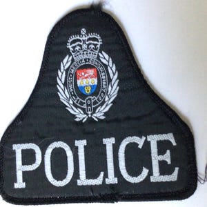 May include: A black, bell-shaped embroidered patch featuring the word POLICE in white capital letters at the bottom. Above the text is a circular emblem with a crown, laurel leaves, and a shield containing red, white, and blue colours.
