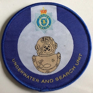 May include: A circular patch with a blue border and the words "UNDERWATER AND SEARCH UNIT". The patch features a diving helmet and the Sussex Police emblem.