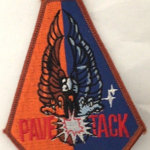 May include: A trapezoidal embroidered patch with a brown border. The background is split vertically into orange and blue halves. A central winged emblem features a white starburst at the bottom. The red text PAVE TACK is stitched across the base.