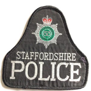 May include: A dark grey embroidered patch bearing the words "STAFFORDSHIRE POLICE" in white. The patch features a detailed emblem with a crown, a starburst design, and a green and white circular element.