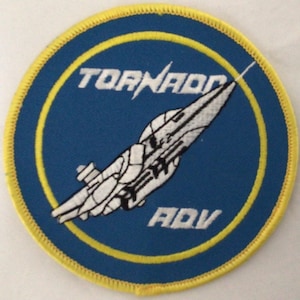 May include: A circular embroidered patch with a blue background and a yellow border. It features a white graphic of a fighter jet angled diagonally. The text TORNADO is embroidered in white above the jet, and the letters ADV are below the jet.