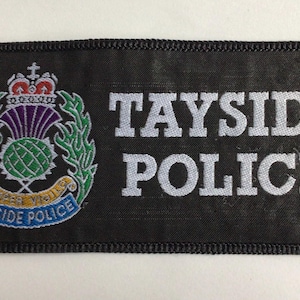 May include: A rectangular black embroidered patch featuring the text TAYSIDE POLICE in white capital letters. To the left is a circular emblem displaying a green thistle, a crown, and green leaves, with a blue banner reading TAYSIDE POLICE.