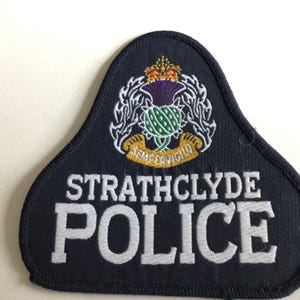 Puede incluir: Parche azul marino de la policía de Strathclyde con texto bordado en blanco y un emblema detallado con un cardo, una corona y una pancarta. El parche tiene una parte superior curvada y una parte inferior puntiaguda.