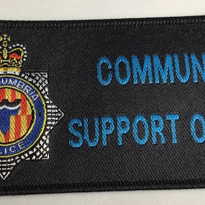 Puede incluir: Un parche rectangular negro bordado que presenta el emblema de la policía de Northumbria a la izquierda, que incluye una corona, una estrella y un escudo central con rayas rojas y amarillas. El texto COMMUNITY SUPPORT OFFICER está bordado en azul.