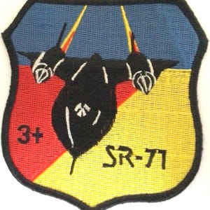 Könnte beinhalten: Gestickter Aufnäher mit einem schwarzen SR-71-Flugzeugdesign. Der Aufnäher hat eine Schildform mit blauem Oberteil, roter linker Seite und gelber rechter Seite. Der Text "3+" und "SR-71" sind sichtbar.