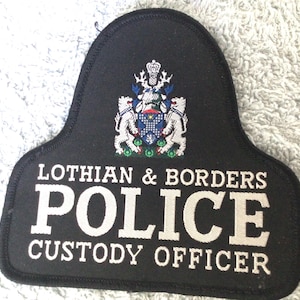 May include: Black patch with white text that reads "LOTHIAN & BORDERS POLICE CUSTODY OFFICER." The patch has a crest at the top with two lions and a crown. The patch is a shield shape.
