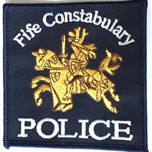 May include: A navy blue square patch featuring the words "Fife Constabulary POLICE" in white embroidery. A gold embroidered image of a knight on horseback is centered on the patch.