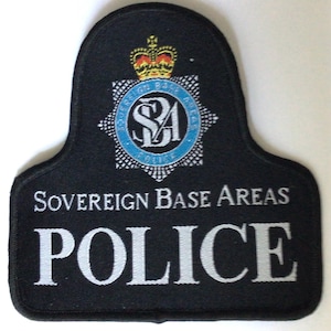 May include: A black police patch with white embroidered text reading "Sovereign Base Areas POLICE." The patch features a blue and silver emblem with a crown at the top.