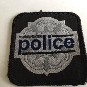 Può includere: Una toppa quadrata ricamata con bordo nero che presenta un emblema di rosa Tudor grigia al centro. Il testo Northamptonshire è stampato in piccole lettere nere sopra la parola police, scritta in grandi lettere blu scuro.