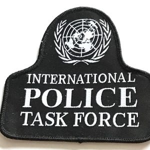 May include: Black patch with white text and a white emblem. The text reads "INTERNATIONAL POLICE TASK FORCE". The emblem is a globe surrounded by a wreath. The patch has a black border.