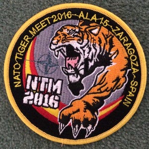 Könnte beinhalten: Ein kreisförmiger gestickter Aufnäher mit einem brüllenden Tiger mit orangefarbenen und schwarzen Streifen. Der Text lautet NATO TIGER MEET 2016 - ALA 15 - ZARAGOZA - SPAIN und NTM 2016. Das Design umfasst einen gelben Rand, einen grauen Hintergrund.