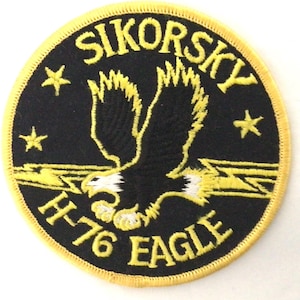 Può includere: Una toppa ricamata circolare con sfondo nero e bordo giallo. Presenta una grafica centrale con un'aquila, fulmini gialli e tre stelle. Il testo riporta SIKORSKY in alto e H-76 EAGLE in basso in colore giallo.
