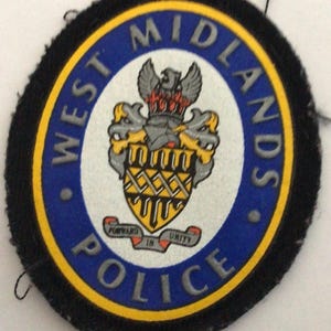 Può includere: Toppa circolare della polizia di West Midlands. La toppa ha un bordo blu e giallo e le parole "WEST MIDLANDS POLICE". L'emblema centrale è uno stemma con una corona e un'aquila.