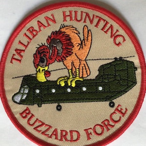 May include: A circular patch with a red border and tan center. The patch features a cartoon vulture perched on a green helicopter. The text "TALIBAN HUNTING" and "BUZZARD FORCE" are in red, arched around the top and bottom of the patch.
