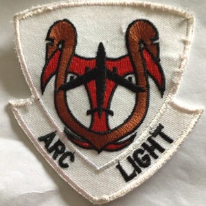 May include: A shield-shaped embroidered patch with a white background and a stitched border. The design features a central black airplane silhouette over a red shape, surrounded by a brown horseshoe-like element. The text ARC LIGHT is in black.