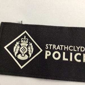 Könnte beinhalten: Ein schwarzer Strathclyde Police-Aufnäher mit weißer Schrift und einem weißen Emblem. Das Emblem ist ein Diamant mit einem Kronen- und Distel-Design. Der Aufnäher ist rechteckig.
