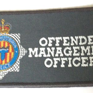 以下が含まれることがあります： 「OFFENDER MANAGEMENT OFFICER」の白い文字が入った長方形のパッチ。王冠と「NORTHUMBRIA POLICE」の文字が入ったバッジも付いています。パッチは濃いグレーで、茶色の縁取りがあります。