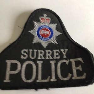 Puede incluir: Un parche bordado negro con el texto SURREY POLICE en hilo plateado. Sobre el texto hay un emblema de estrella plateada con un centro circular rojo, blanco y azul coronado por un pequeño diseño de corona roja y blanca sobre fondo negro.