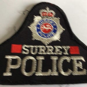 Puede incluir: Un parche bordado negro que presenta el emblema de la policía de Surrey en la parte superior, que incluye una estrella plateada, un círculo azul y un centro rojo con una corona. El texto SURREY POLICE está bordado en hilo plateado.