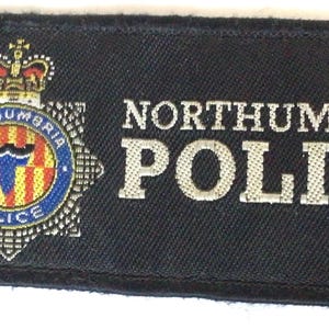 May include: A rectangular black patch with the words "NORTHUMBRIA POLICE" in white lettering. The patch features a detailed emblem with a crown, a shield, and the words "NORTHUMBRIA POLICE" in a circular design.