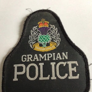 Könnte beinhalten: Ein schwarzer, glockenförmiger bestickter Aufnäher mit dem Text GRAMPIAN POLICE in weißen Großbuchstaben. Über dem Text befindet sich ein farbiges Emblem einer violett-grünen Distel mit einer Krone und einem gelben Banner mit der Aufschrift SEMPER VIGILO.