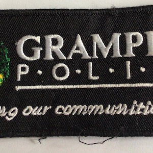 May include: A rectangular black embroidered patch featuring the text GRAMPIAN POLICE in white capital letters above a horizontal line and the phrase keeping our communities safe below. To the left is a colorful emblem with a crown and thistle.