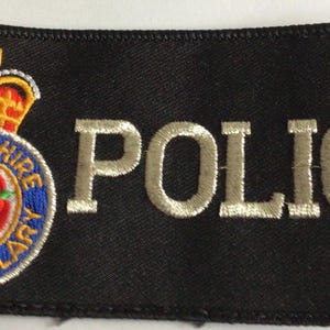 May include: A rectangular black embroidered patch featuring the word POLICE in silver thread. To the left is a circular emblem with a red rose, blue border, and gold crown, with the text LANCASHIRE CONSTABULARY encircling the central rose.