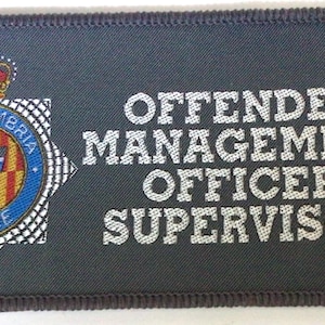 May include: A grey rectangular patch with the text "OFFENDER MANAGEMENT OFFICER SUPERVISOR" in white. It features the Northumbria Police badge, a crown, and a blue, red, and yellow emblem. The patch has a stitched border.