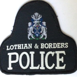 May include: A black police shoulder patch with white embroidered text that reads "LOTHIAN & BORDERS POLICE". The patch features a detailed emblem with a crest, two lions, and a blue and white shield.