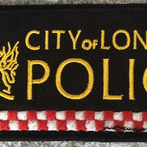 May include: Black rectangular patch with gold embroidered text "CITY of LONDON POLICE". Features a gold emblem with a red and white shield. A red and white checkered pattern is at the bottom.