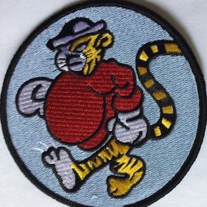 May include: Embroidered patch featuring a cartoon tiger character in a red shirt and a purple hat, with a yellow and black striped tail. The patch is circular, with a light blue background and a black border.