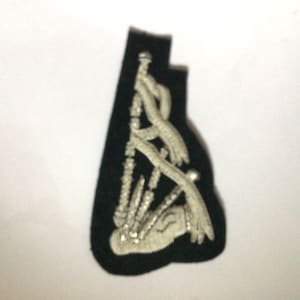 May include: Black embroidered patch with a white and silver bagpipe design. The patch is shaped uniquely and is intended for sewing onto garments or accessories. The design features intricate stitching details.