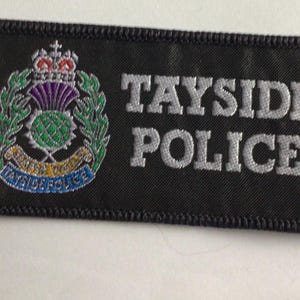 May include: A rectangular black embroidered patch featuring the text TAYSIDE POLICE in silver thread. To the left is a colorful emblem depicting a purple and green thistle, a silver crown, and a blue banner with the text TAYSIDE POLICE.