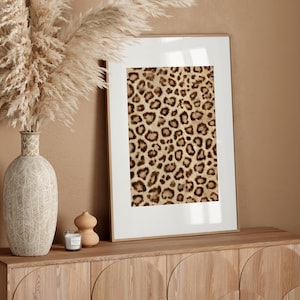 May include: A framed leopard print art piece with a white border rests on a wooden cabinet. A vase with dried pampas grass and a candle sit nearby. The artwork features a brown and tan animal print.