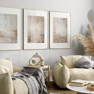 May include: Three framed abstract paintings in muted beige and brown tones hang on a light grey wall above two beige armchairs with grey throw pillows. A small wooden side table with a ceramic vase and a candle sits between the chairs. A vase with dried pampas grass sits on a white marble coffee table with a book and a tray.