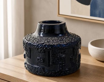 May include: A dark navy blue ceramic vase with a wide, cylindrical body and a narrow neck. The exterior features a textured, pebbled surface finish combined with a repeating geometric pattern embossed around the center of the vessel.