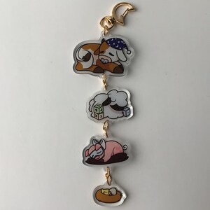May include: A gold chain keychain with four charms. The charms are a sleeping cow wearing a blue and white star hat, a sleeping sheep with a green, yellow, and blue block, a sleeping pig wearing a pink bow, and a yellow chick in a nest.